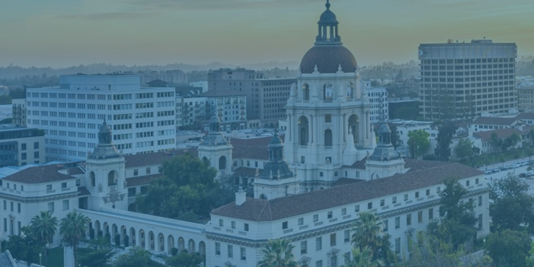 Aerial view of Pasadena City Hall with its dome, surrounded by palm trees and nearby office buildings, under a blue sky at sunset.