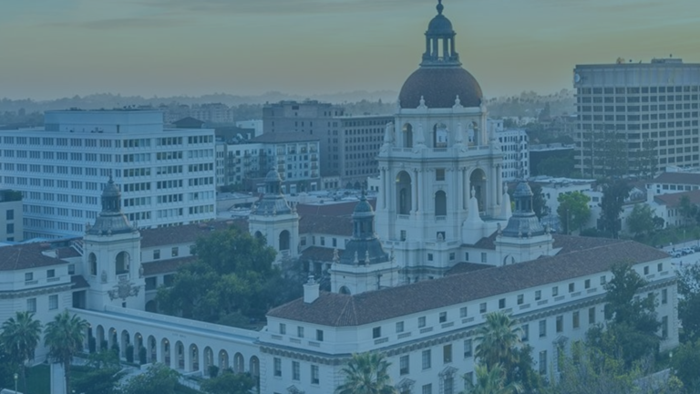 Aerial view of Pasadena City Hall with its dome, surrounded by palm trees and nearby office buildings, under a blue sky at sunset.