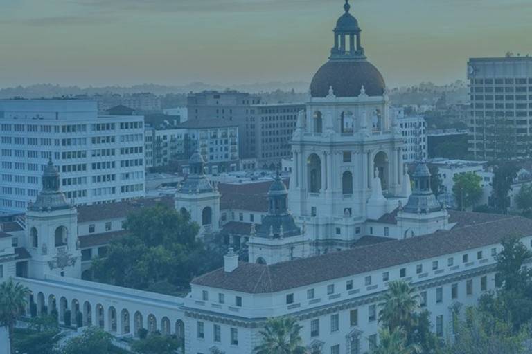 Aerial view of Pasadena City Hall with its dome, surrounded by palm trees and nearby office buildings, under a blue sky at sunset.