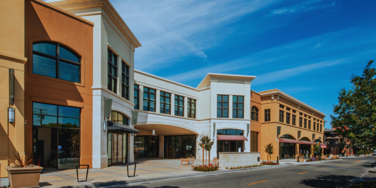 A street-level view of a modern multi-story shopping complex with tan and white stucco storefronts under a clear blue sky.