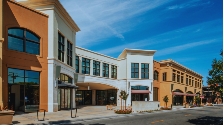 A street-level view of a modern multi-story shopping complex with tan and white stucco storefronts under a clear blue sky.