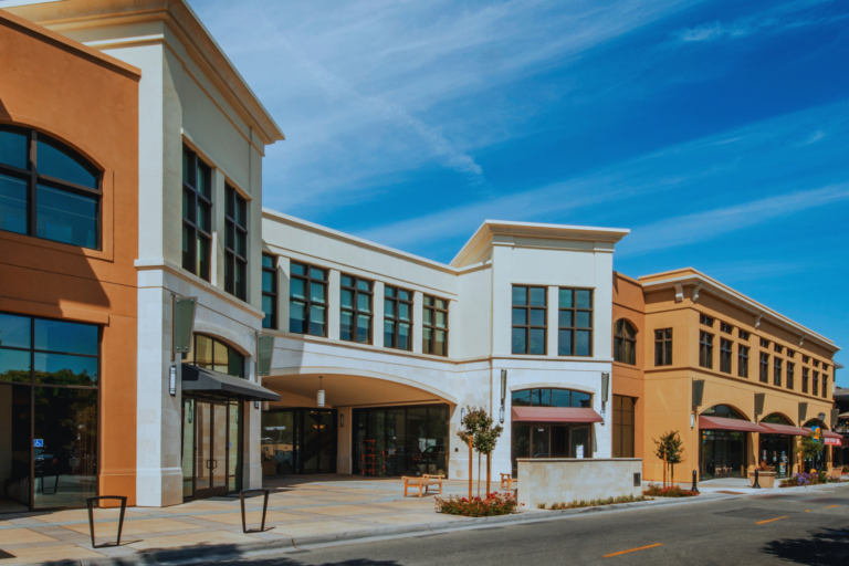 A street-level view of a modern multi-story shopping complex with tan and white stucco storefronts under a clear blue sky.